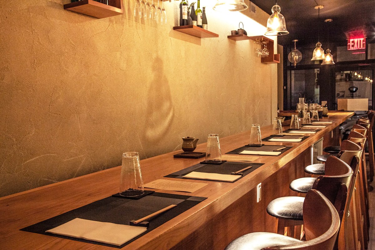 Wooden sushi-style counter with place settings, chopsticks, glasses and wine bottles on shelves, dim warm lighting