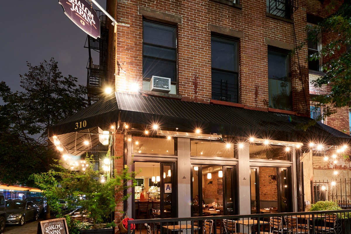 Exterior of Shalom Japan restaurant at night, brick building with awning, string lights and terrace seating