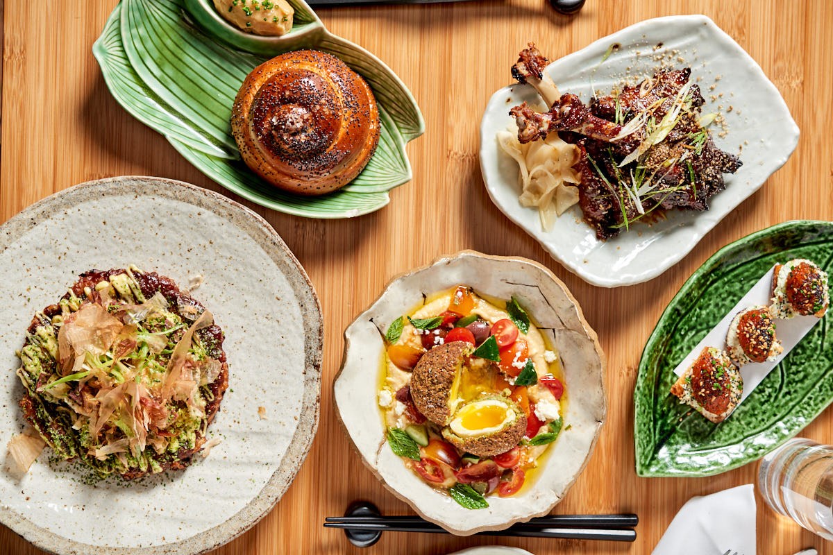 Overhead view of an assortment of dishes including challah bread, ribs, okonomiyaki and tuna tartare on wooden table