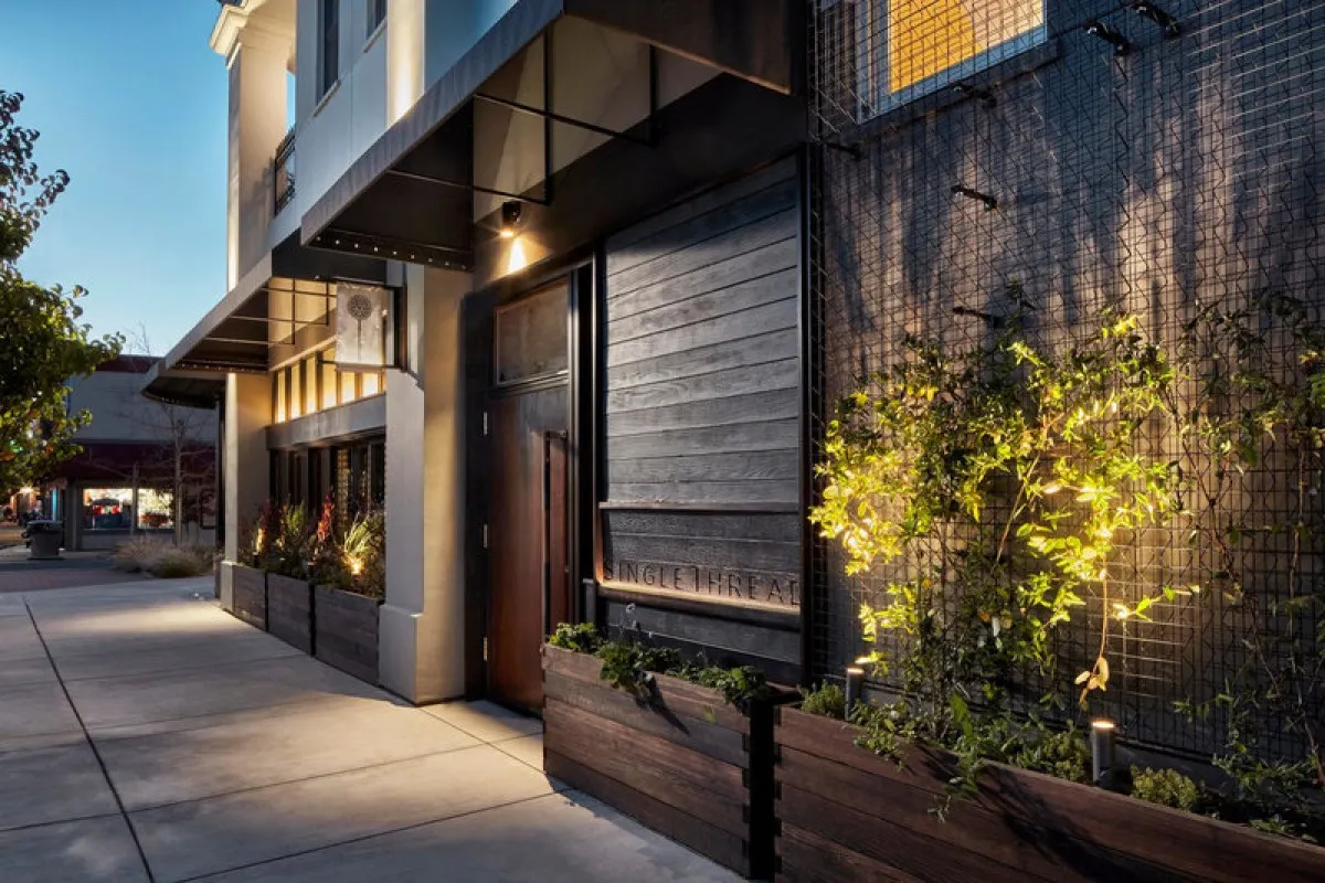Evening exterior of SingleThread Inn entrance with wooden door, illuminated signage, and lush planters along a sidewalk