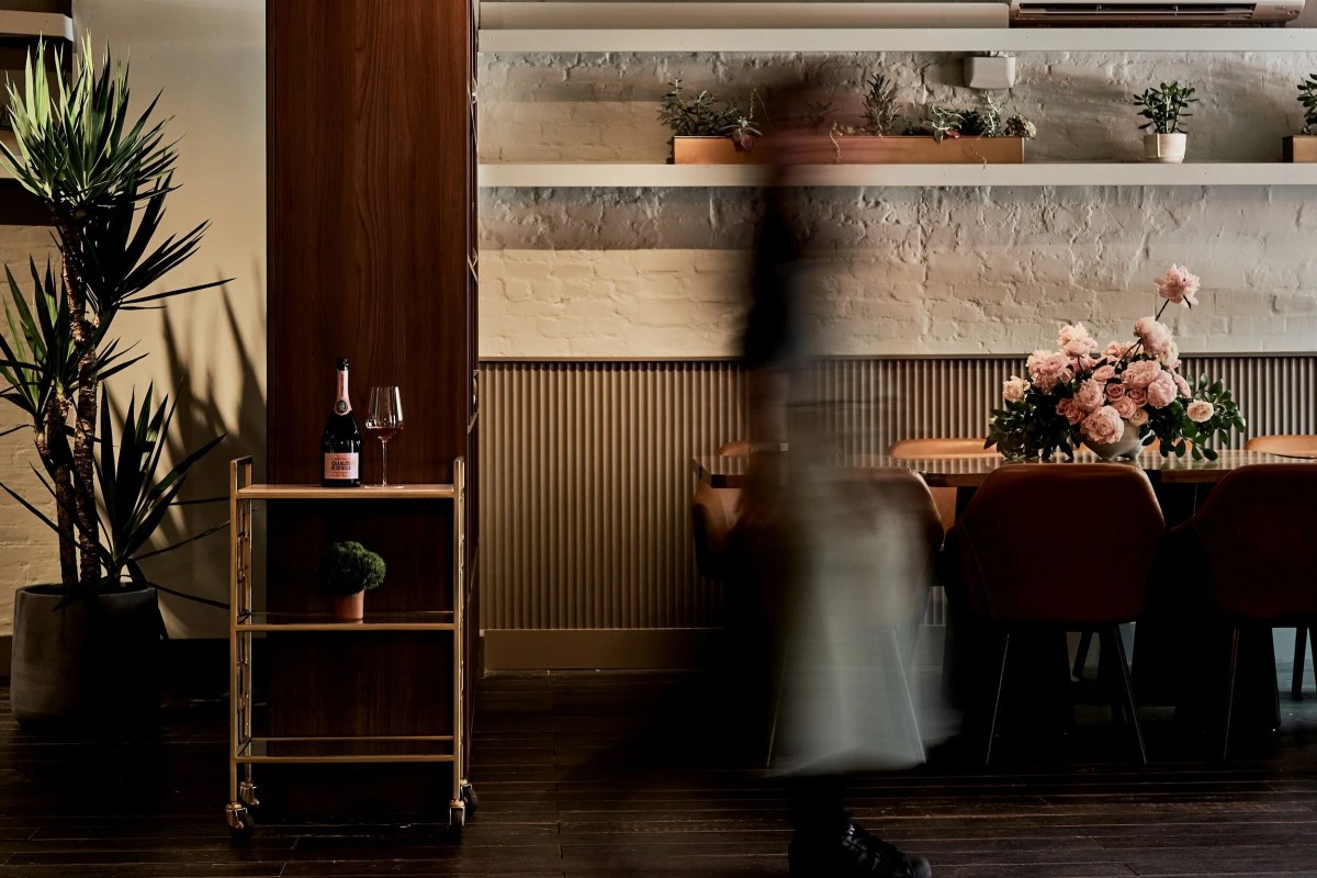Dining room with plants, wine bottle on cart, blurred figure walking past floral table setting