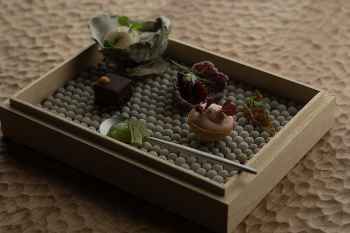 A wooden tray filled with small stones holding several delicate bite-sized dishes including an oyster shell, chocolate cube, and tart