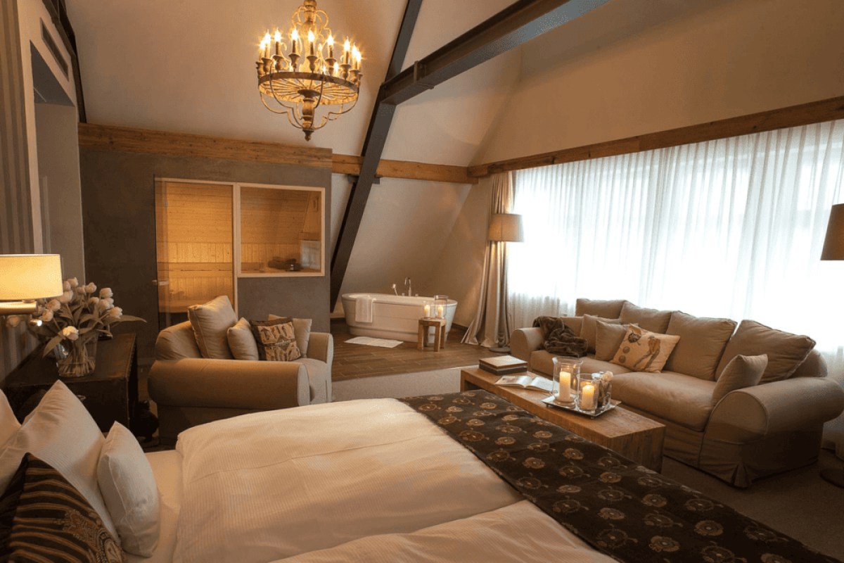 Luxurious hotel suite with four-poster-style bed, sitting area, chandelier, and a freestanding bathtub under sloped wooden-beamed ceiling.