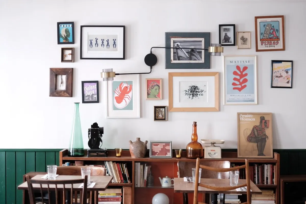 Dining room wall with eclectic gallery of framed art, posters, and vintage objects on a wooden bookshelf below
