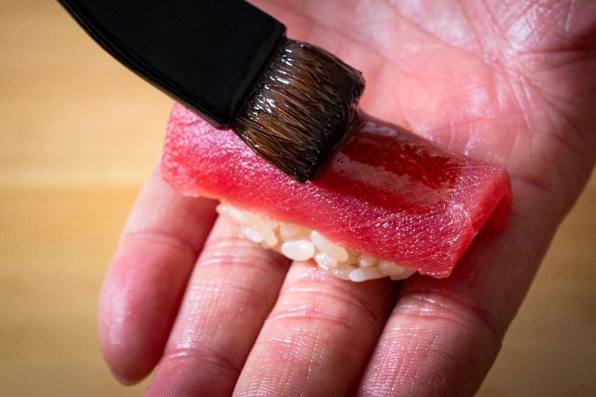 Hand holding a piece of nigiri sushi with tuna being brushed with sauce
