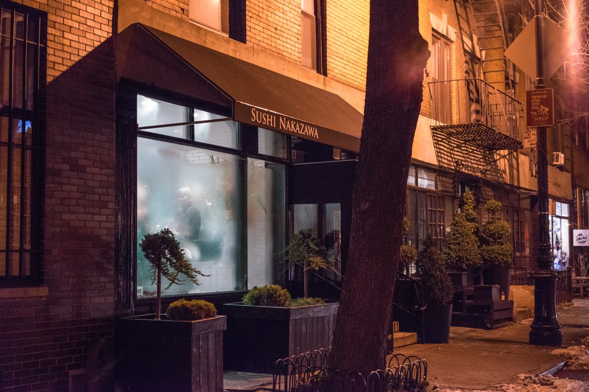 Night view of Sushi Nakazawa restaurant exterior with illuminated sign, awning, and potted plants on sidewalk