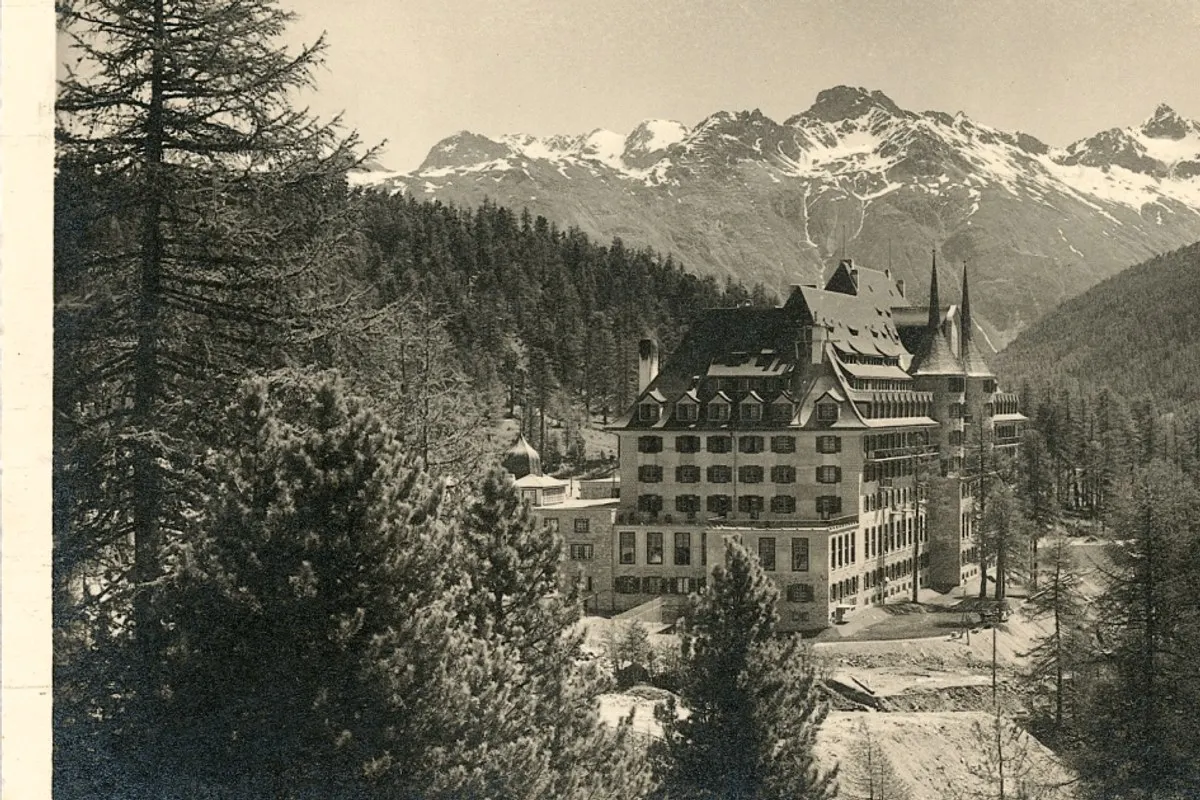 Vintage black-and-white photo of Suvretta House hotel exterior surrounded by pine forest with snow-capped Alps behind