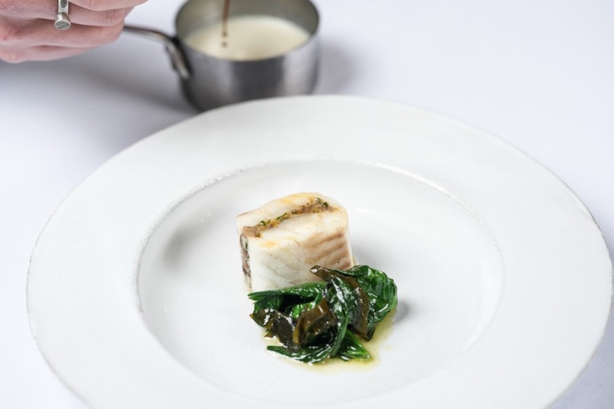 Plated fish dish with greens on white plate, chef's hand pouring sauce from small pot in background