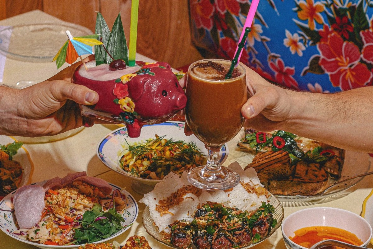 Hands holding a pig-shaped cocktail and a creamy coffee drink over a table spread of Vietnamese dishes