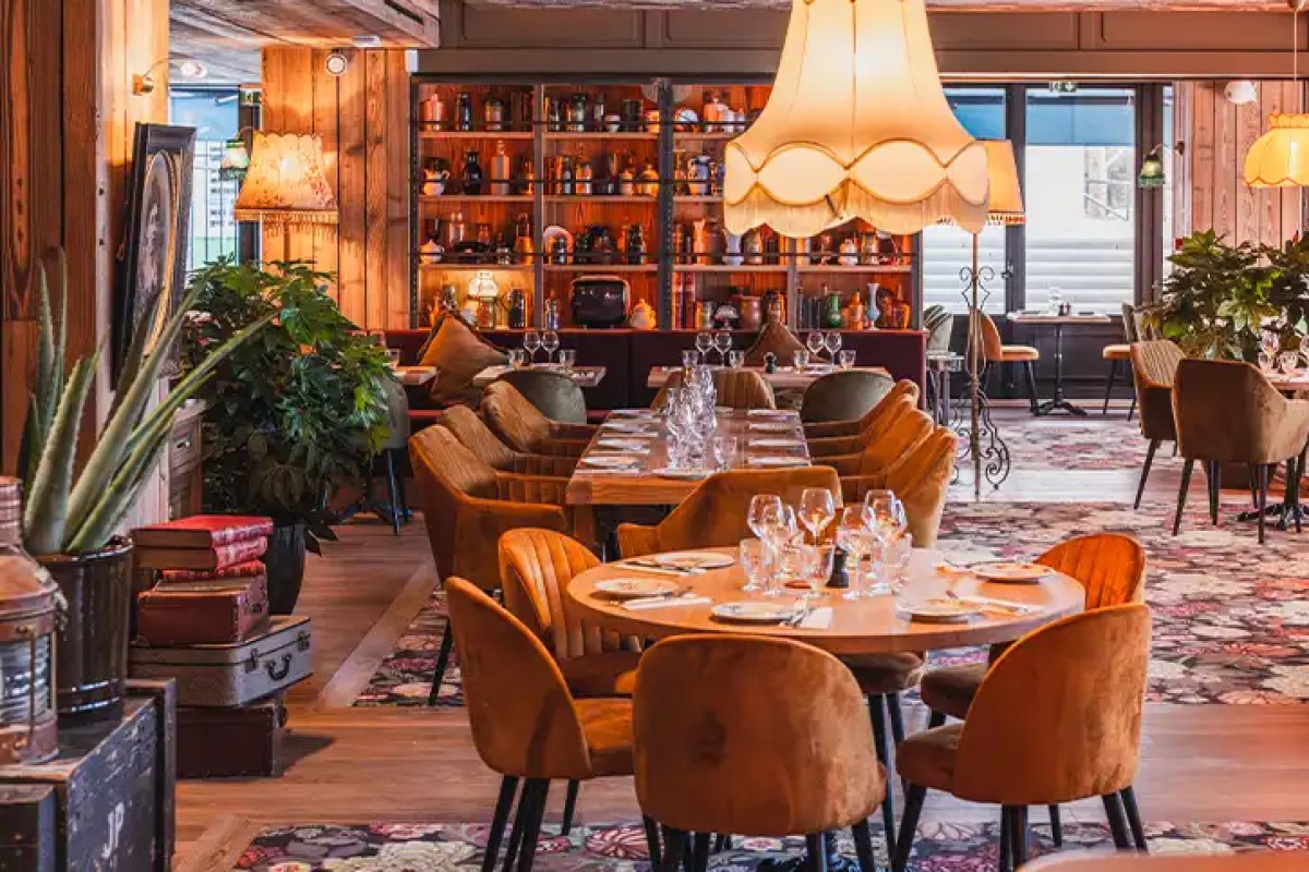 Cozy restaurant dining room with wooden beams, velvet chairs, set tables, pendant lamps with fringed shades, and shelves of decorative objects