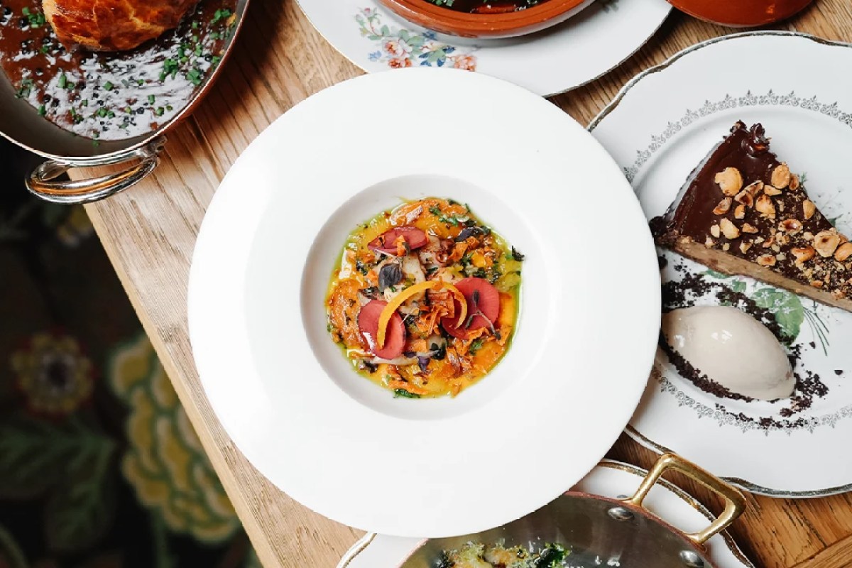 Overhead view of a plated dish with vegetables and seafood on a white plate, surrounded by other dishes and a chocolate hazelnut tart on a wooden table