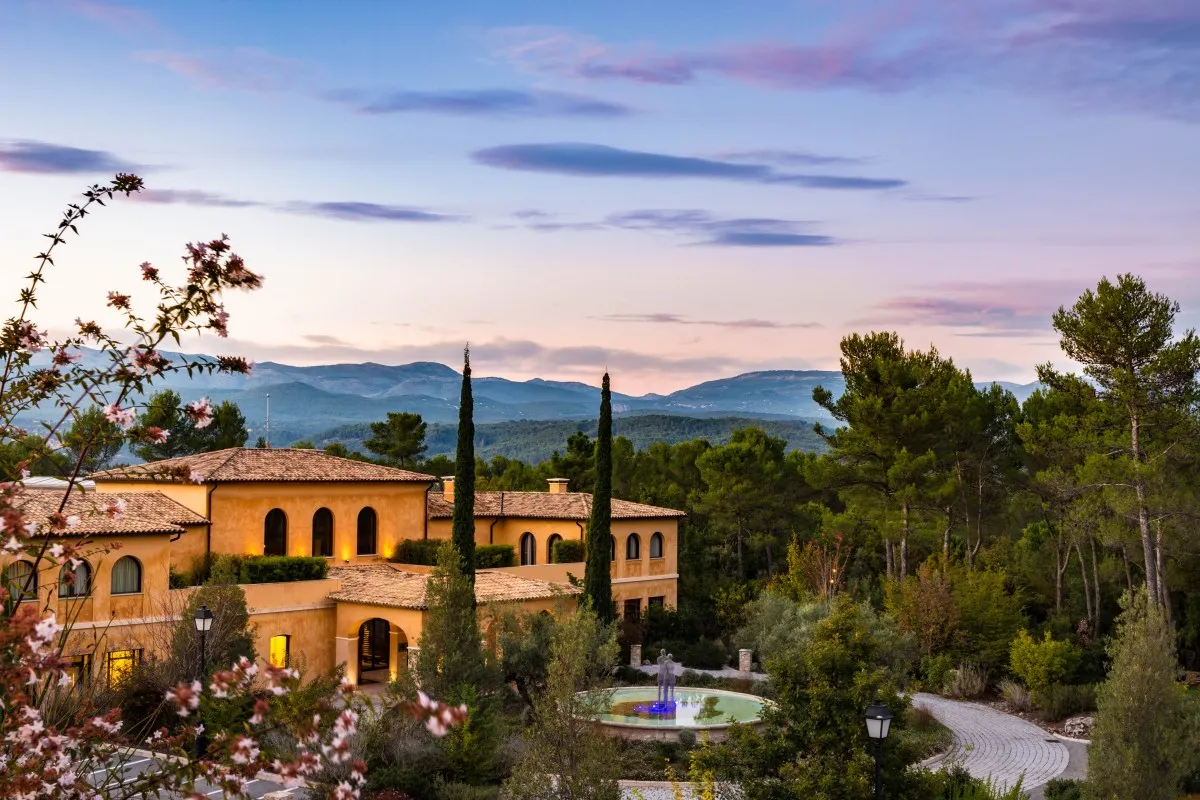 Twilight view of the ochre hotel building with fountain, cypress trees and mountain backdrop