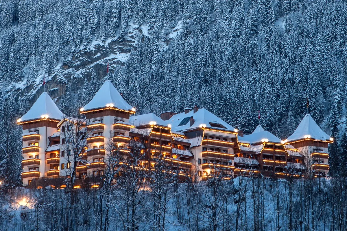 Twilight view of The Alpina Gstaad hotel illuminated with warm lights, surrounded by snowy forest and mountains