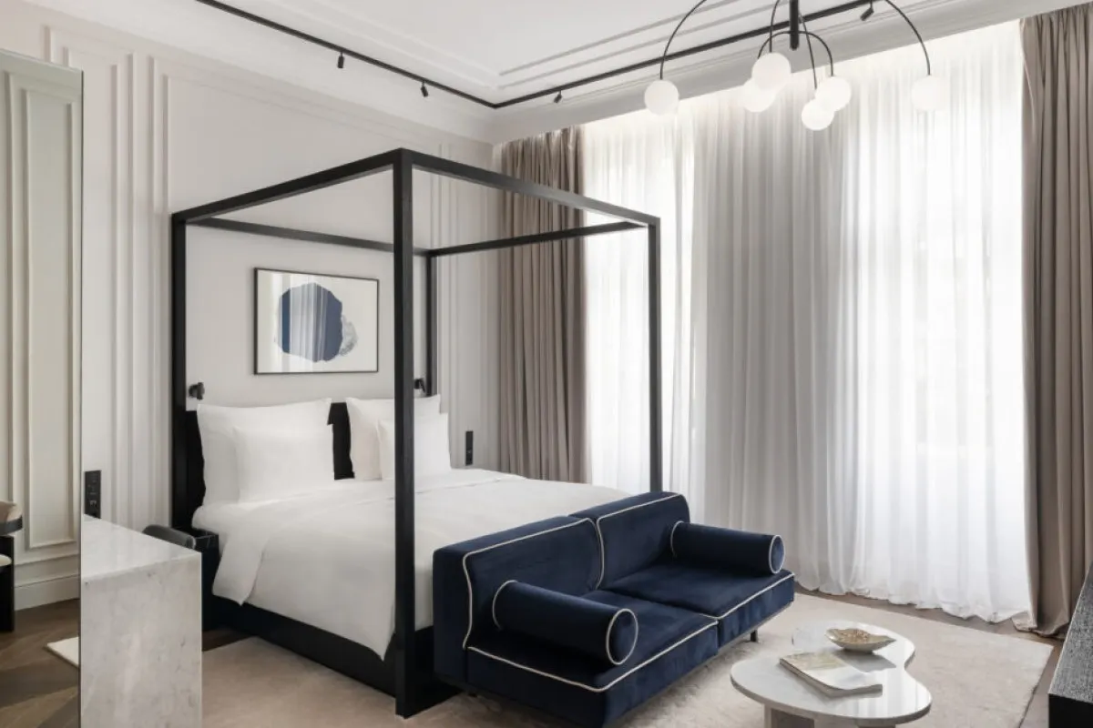 Modern hotel suite with black four-poster bed frame, navy velvet sofa, and tall sheer-curtained windows.
