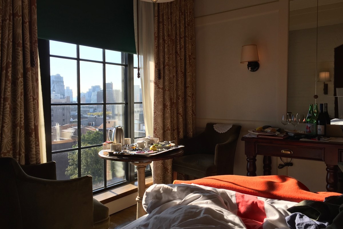 Hotel guest room with unmade bed, breakfast tray by window, armchairs, and city skyline view
