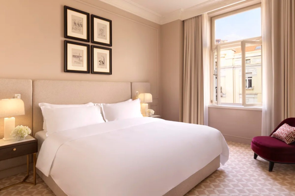 Hotel bedroom with white bedding, framed art, and open window showing historic street view