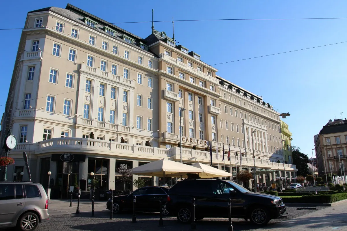 Grand cream-colored hotel facade with 'CARLTON' signage, flags, and awnings under a blue sky, street view.