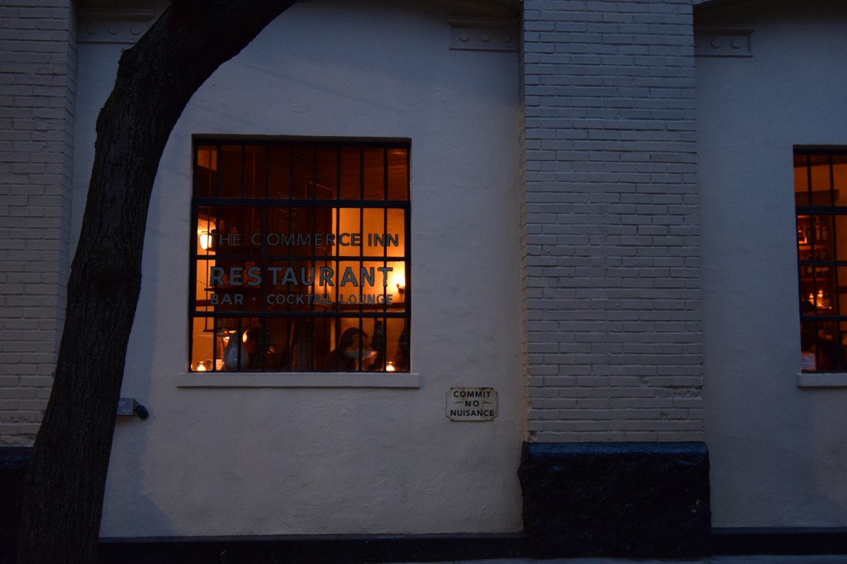Exterior night view of The Commerce Inn restaurant, showing painted brick facade and illuminated window with signage reading 'THE COMMERCE INN RESTAURANT BAR COCKTAIL LOUNGE'.