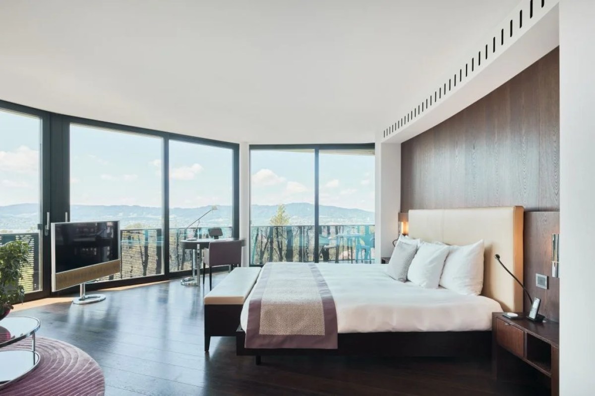 Spacious curved guest room with king bed, wood paneling and panoramic window view of Zurich