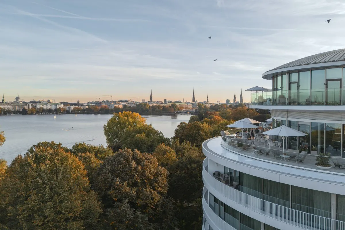 Aerial view of The Fontenay's curved glass façade and rooftop terrace overlooking the Alster lake at sunset, with Hamburg skyline in background.
