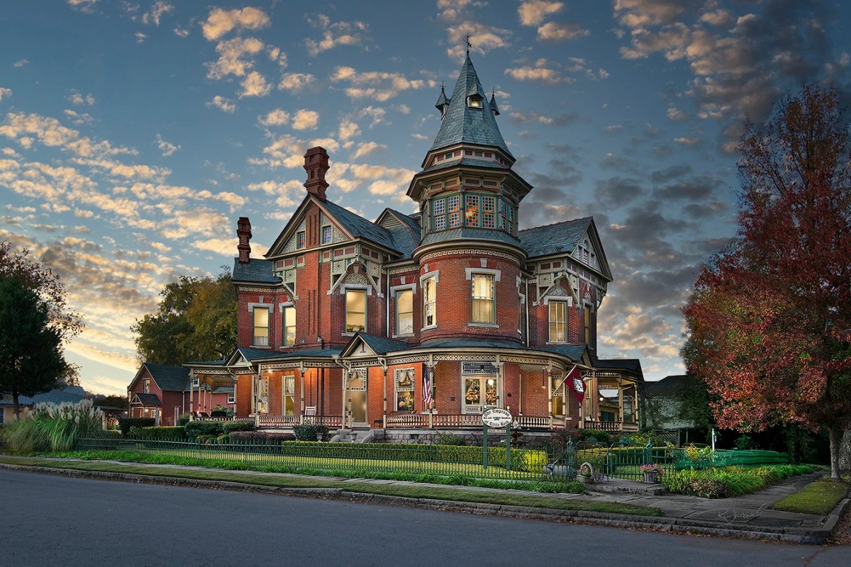 Victorian red-brick mansion with turret facade at sunset, warm sky with clouds, autumn foliage tree beside the building