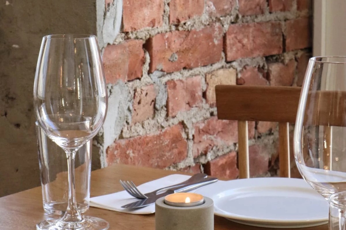 Laid restaurant table with wine glasses, cutlery, white plate, and a lit candle against an exposed brick wall