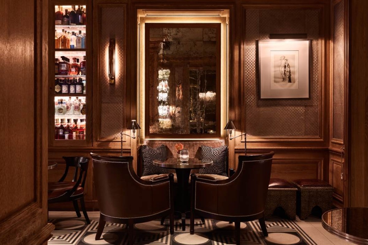 Dark wood-paneled hotel bar with leather chairs, backlit mirror and shelves of spirits