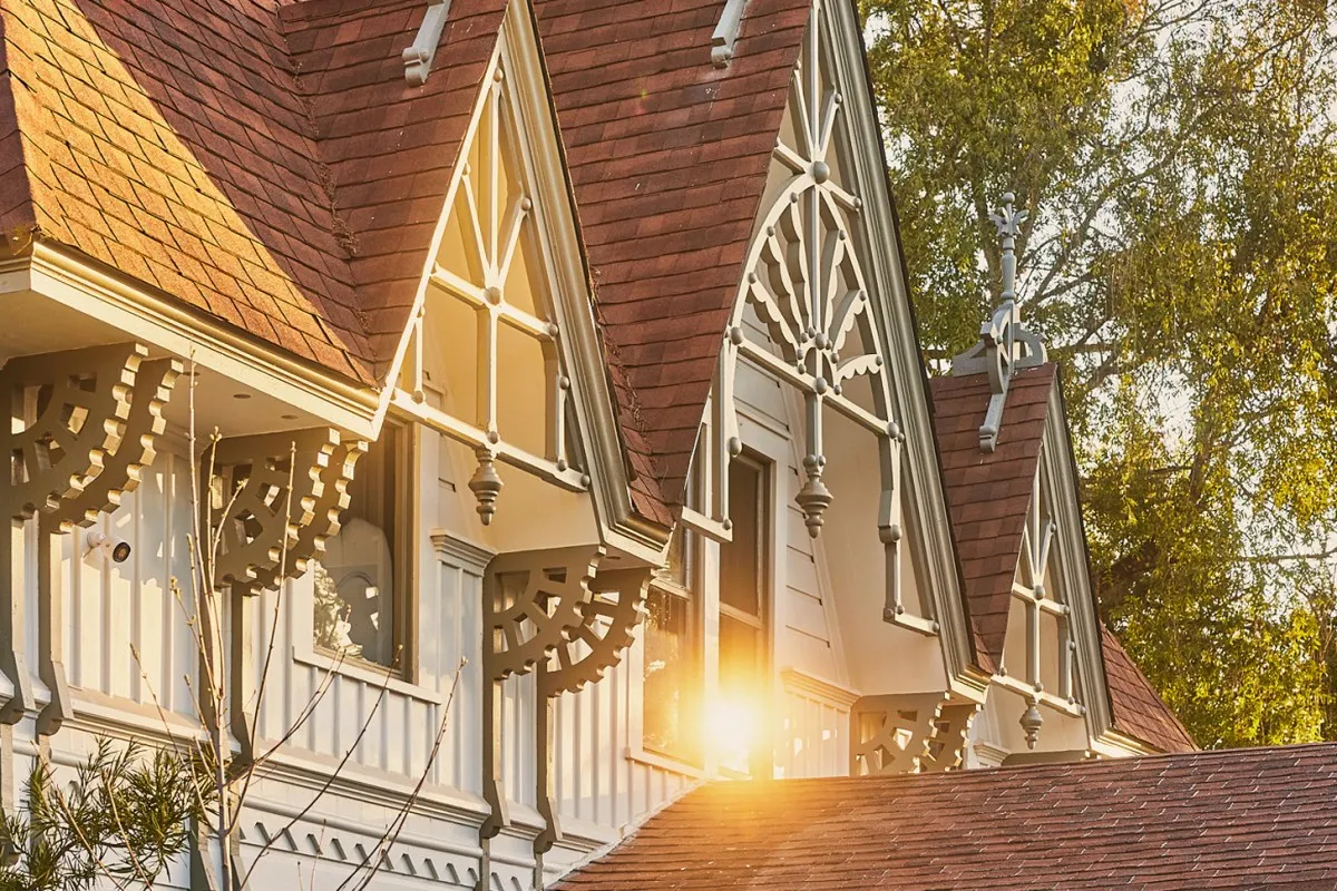 Close-up of Victorian-style roofline with ornate gingerbread trim and gabled dormers lit by golden sunset light, trees in background