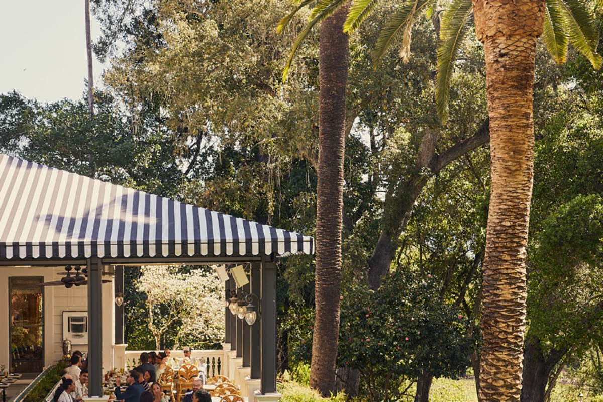 Striped awning dining pavilion with guests seated on terrace surrounded by palms and oak trees