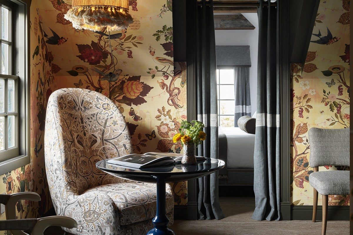 Guest room with floral bird-print wallpaper, patterned armchair, round table, and bed visible through curtains