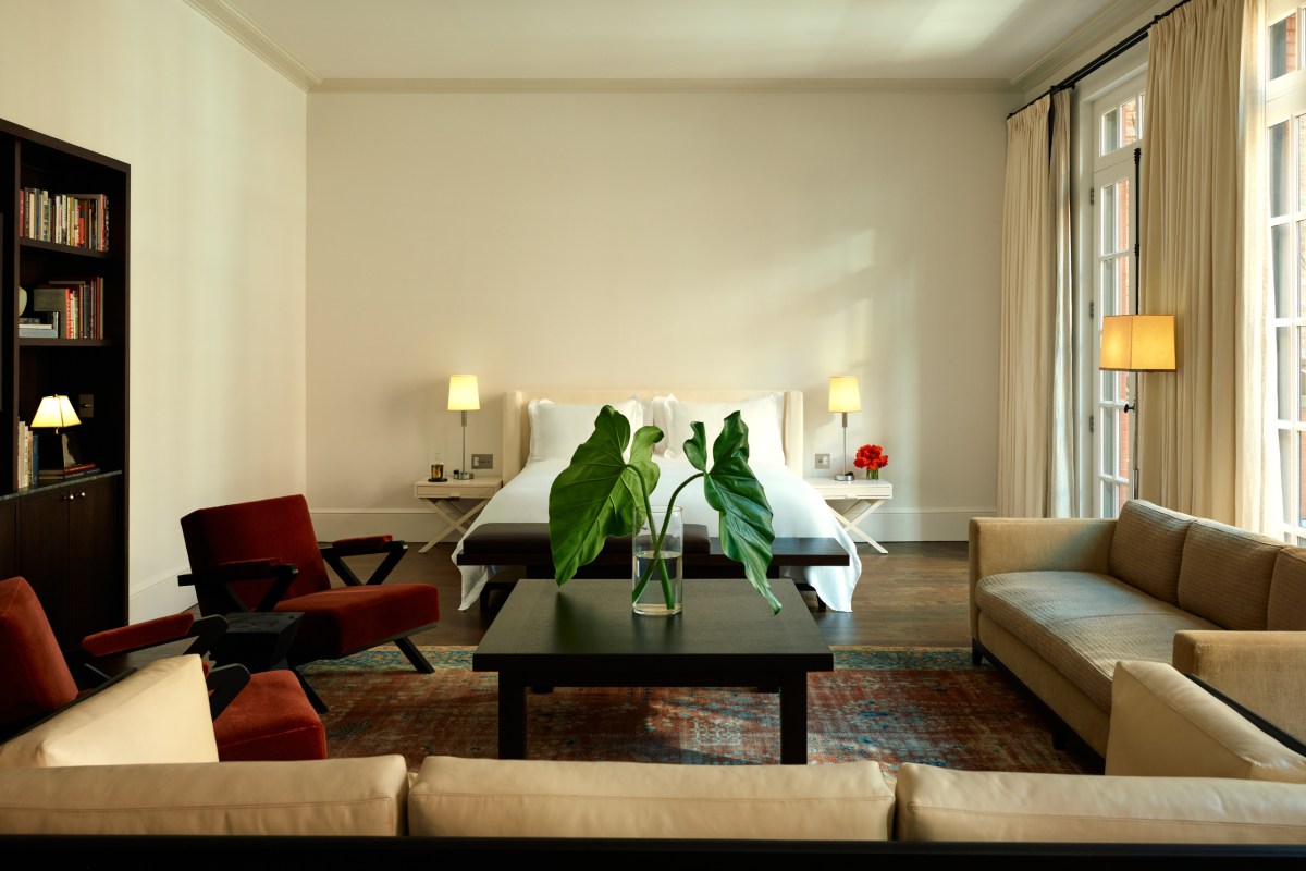 Spacious hotel suite with white bed, red velvet chairs, dark coffee table, and tall curtained windows