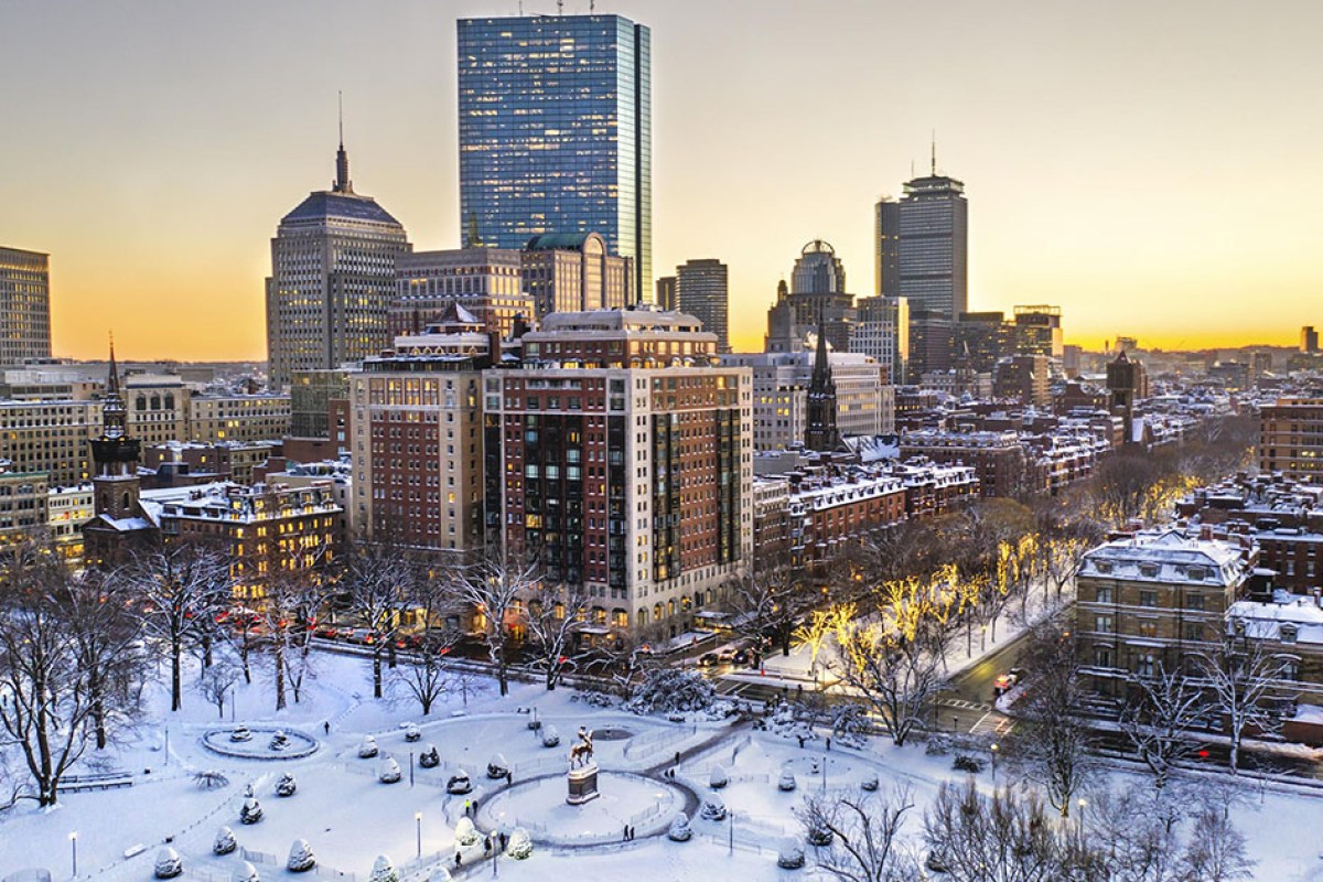 Aerial winter sunset view of Boston skyline with the Newbury Boston's brick and limestone building prominent among city towers, snow-covered park below