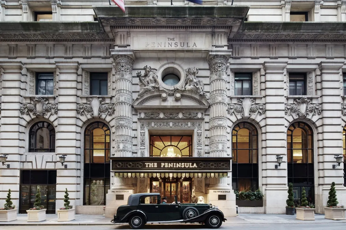 The Peninsula New York hotel façade with vintage Rolls Royce parked in front, ornate stone entrance and gold signage.