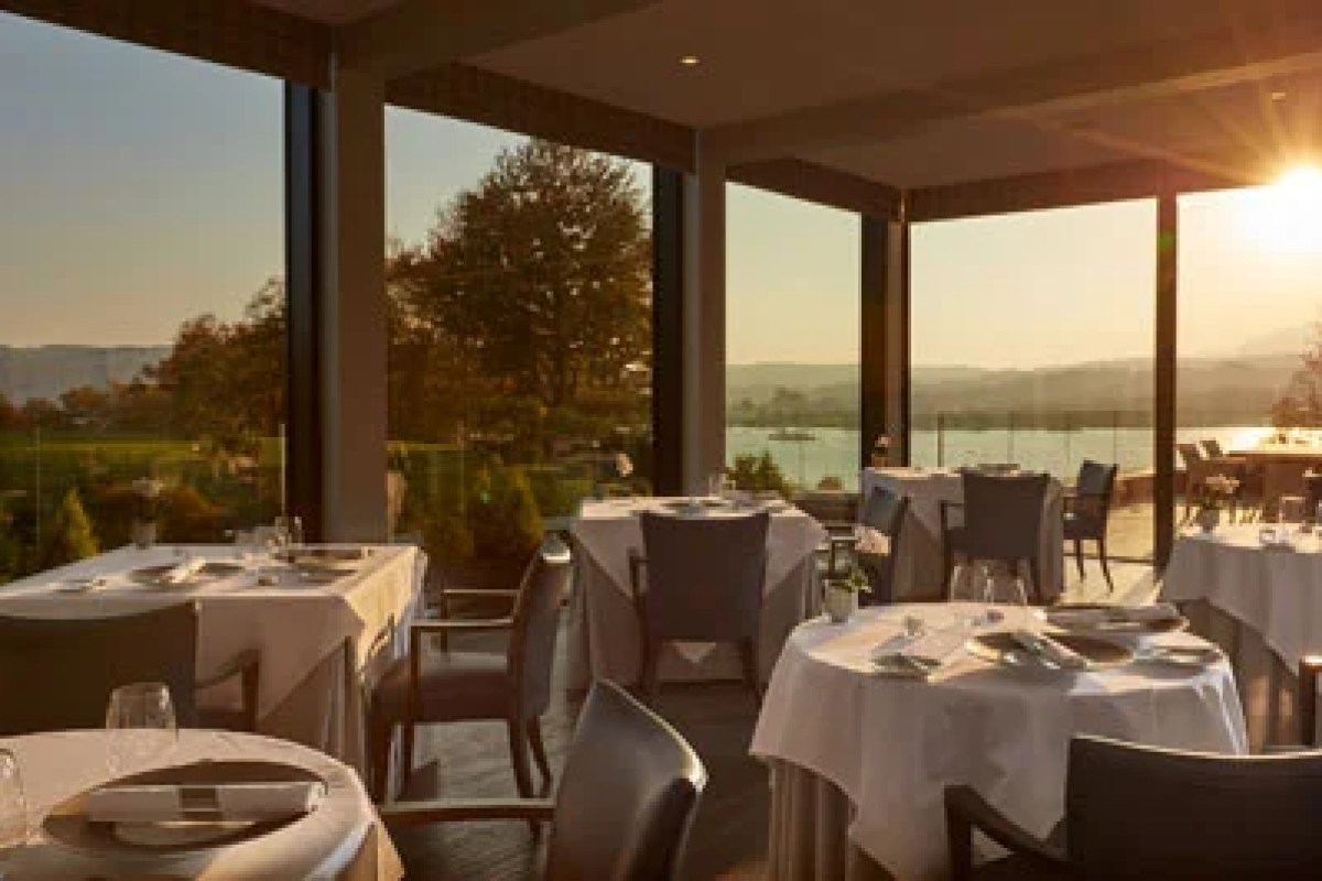 Hotel restaurant with white-clothed tables and floor-to-ceiling windows overlooking a lake at sunset