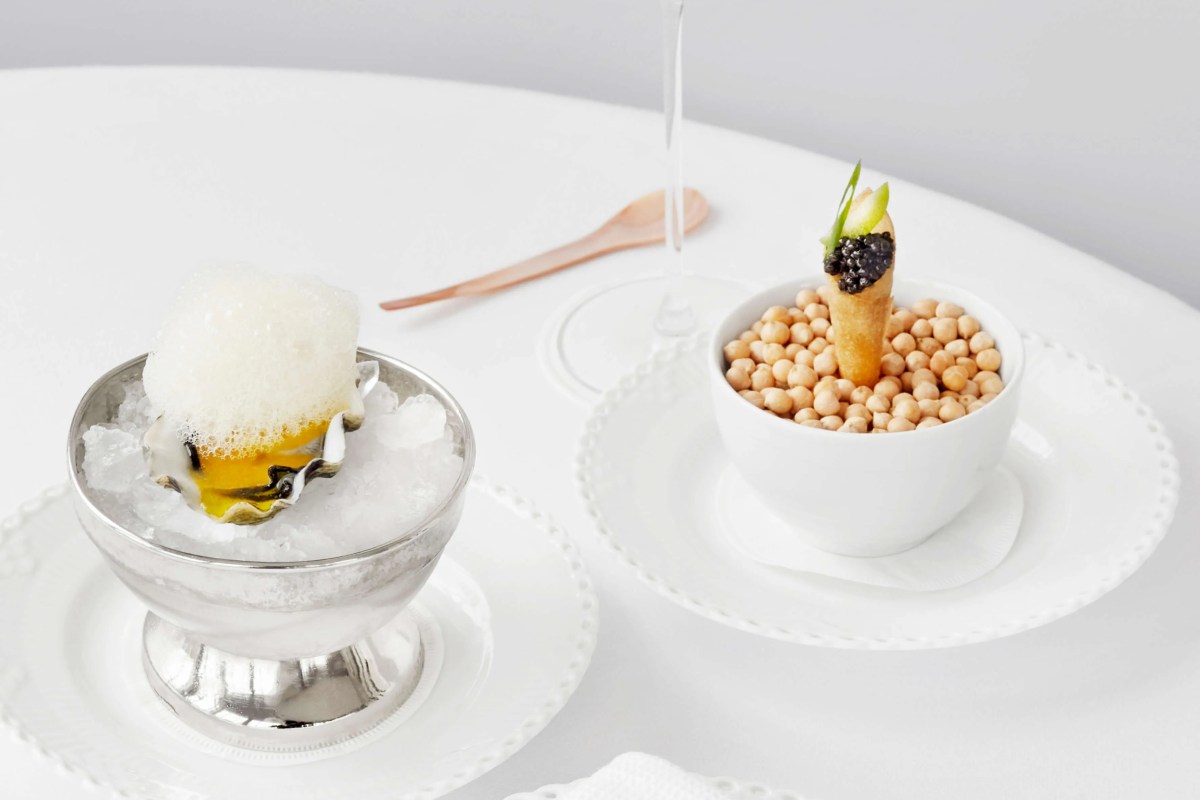 Two plated dishes on white table: an oyster with foam in a silver cup and a caviar-topped tart in a bowl of chickpeas.