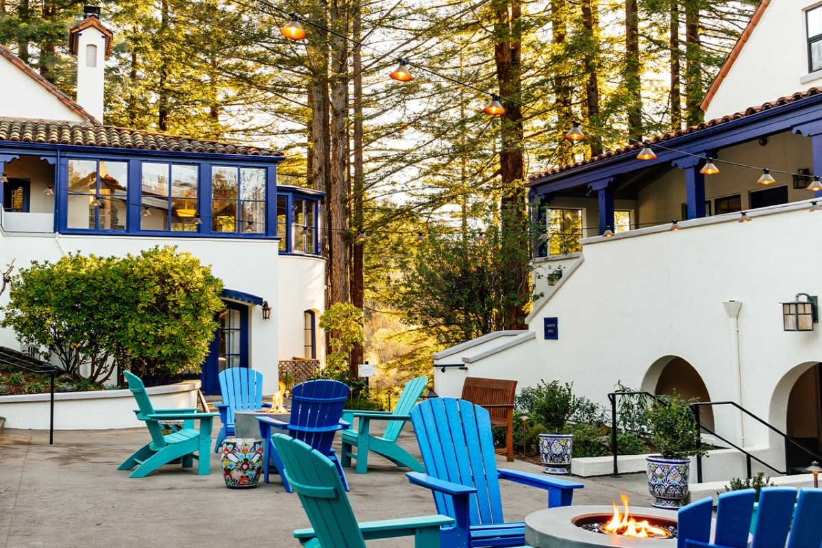Exterior of The Stavrand hotel with blue-trimmed white buildings, string lights, fire pit and colorful Adirondack chairs surrounded by redwood trees