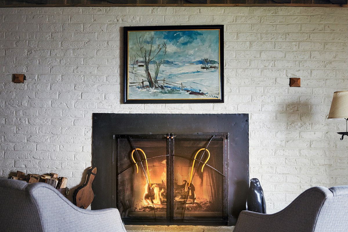 Cozy lounge interior with a lit stone fireplace, armchairs, and a framed landscape painting above the mantel.