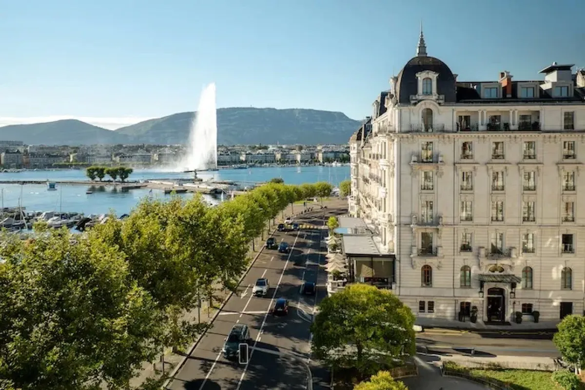Aerial view of The Woodward hotel facade on Geneva lakefront with Jet d'Eau fountain and mountains in background