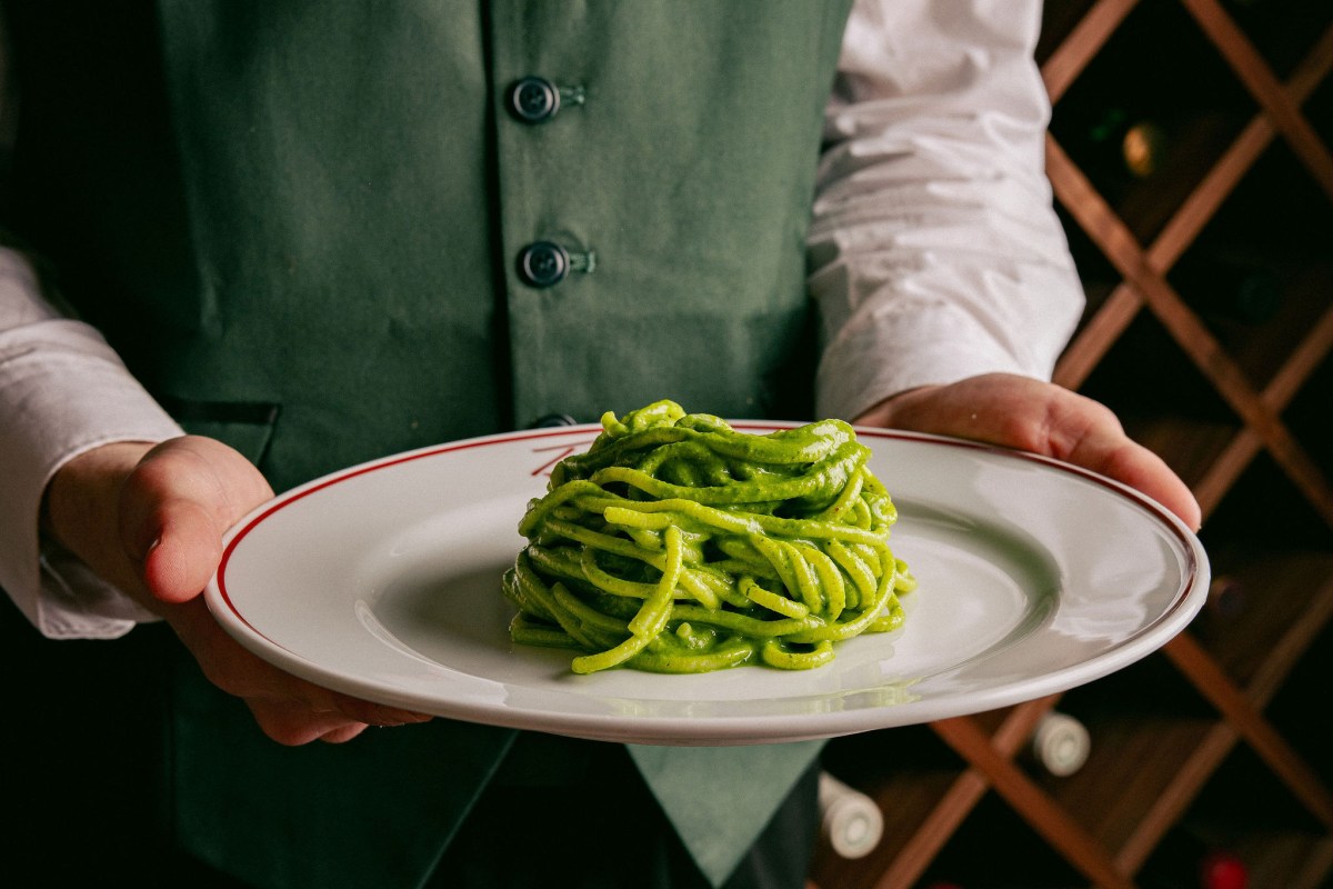 Waiter's hands presenting a plate of green pesto pasta over a wine rack background