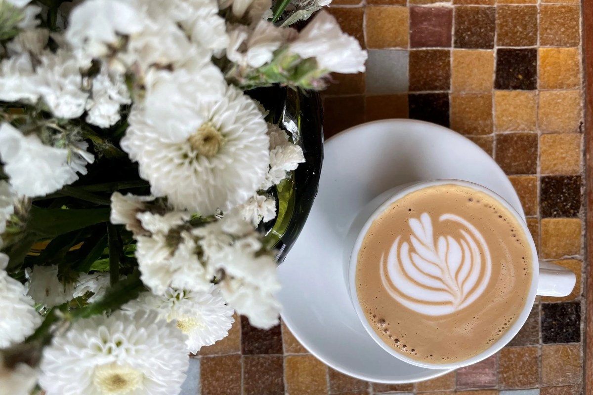 Overhead shot of a latte with leaf-pattern foam art beside white flowers on a tiled table