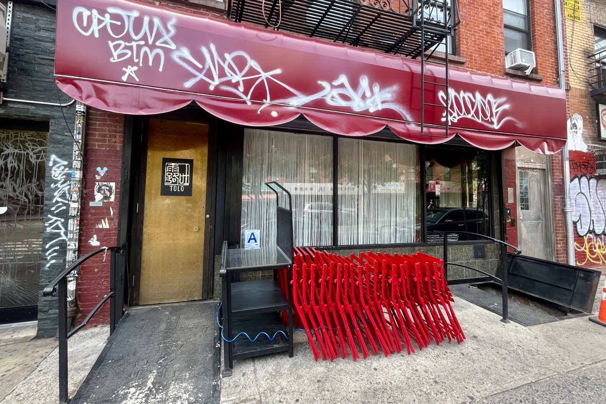 Exterior storefront of Tolo NYC with red awning, gold door, graffiti, and stacked red chairs on sidewalk