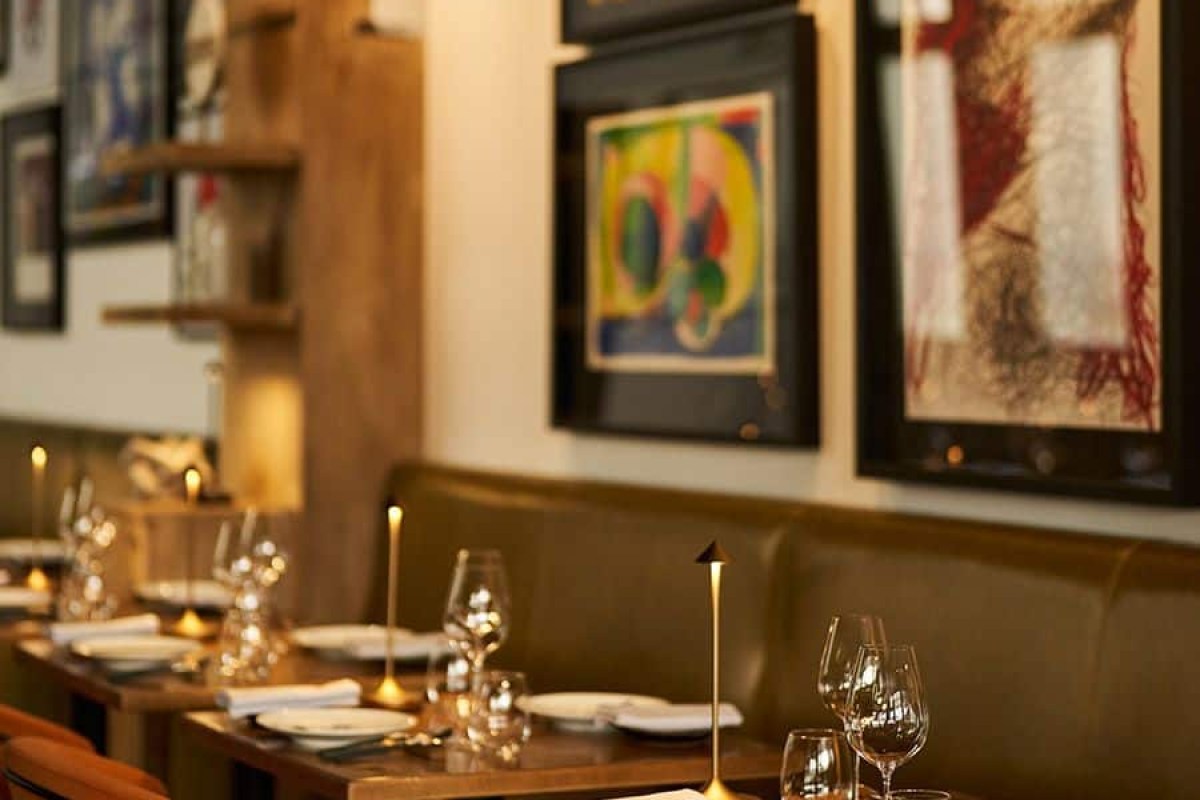 Wide view of dining room with wooden tables set for dinner, leather banquette and gallery wall of art