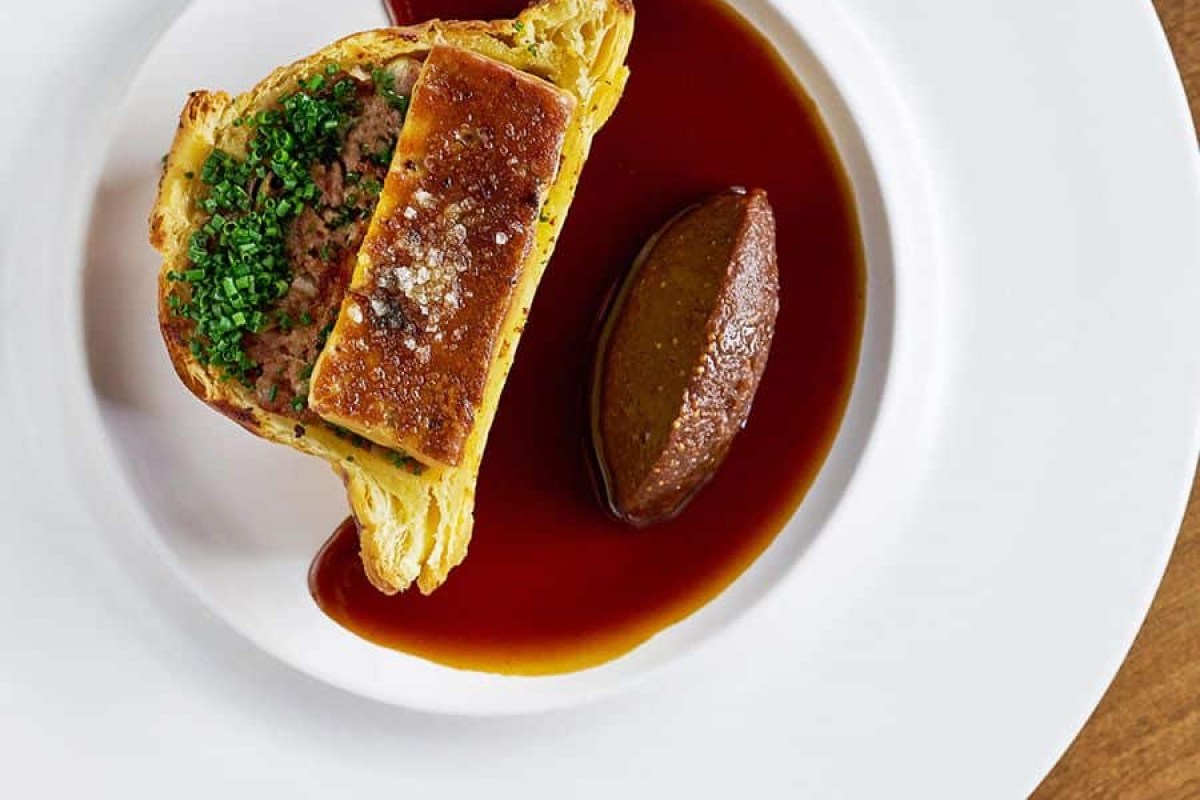 Overhead shot of a pâté en croûte dish with chives and dark sauce on white plate
