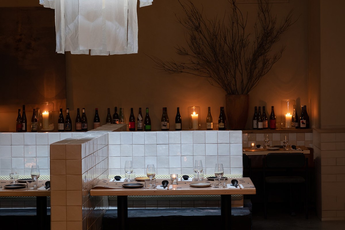 Dimly lit dining room with white-tiled booths, set tables with glasses and candles, and a wine bottle display.