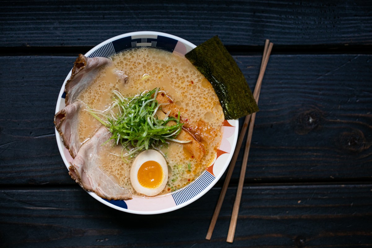 Overhead shot of a tonkotsu ramen bowl with pork slices, scallions, soft-boiled egg, and nori on a dark wood table.