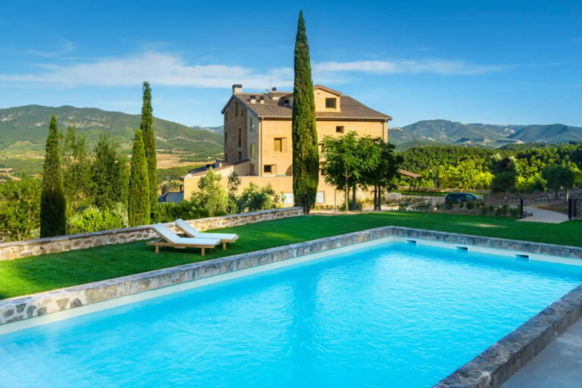 Stone hotel building with cypress trees and mountain views behind an outdoor pool and lawn with loungers