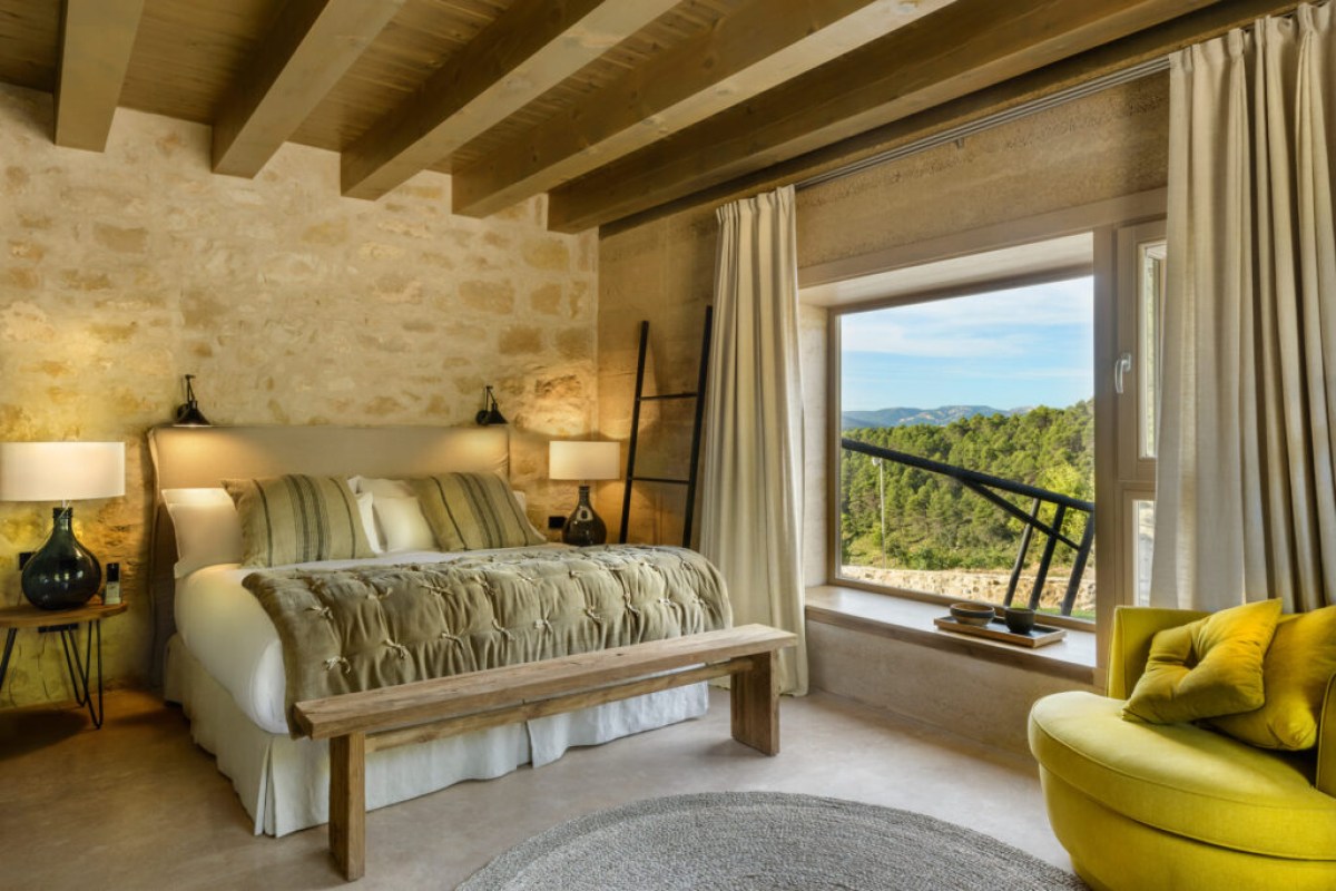 Guest bedroom with stone wall, wooden beams, bench, and window overlooking forested hills