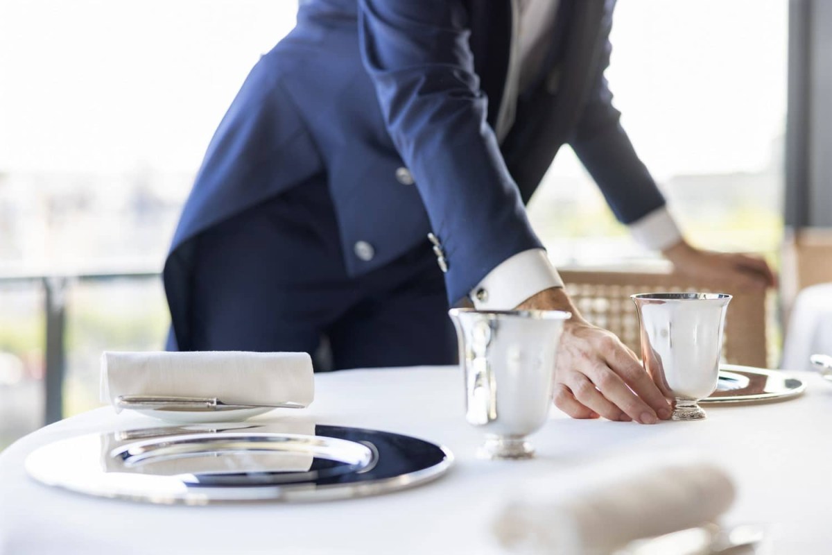 Waiter in dark suit arranging silver cups and folded napkin on a white-clothed table