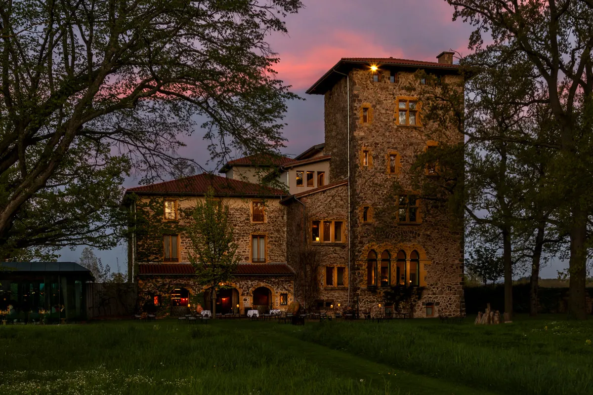 The stone manor house and tower of Troisgros in Ouches at dusk, with the glass restaurant pavilion on the left