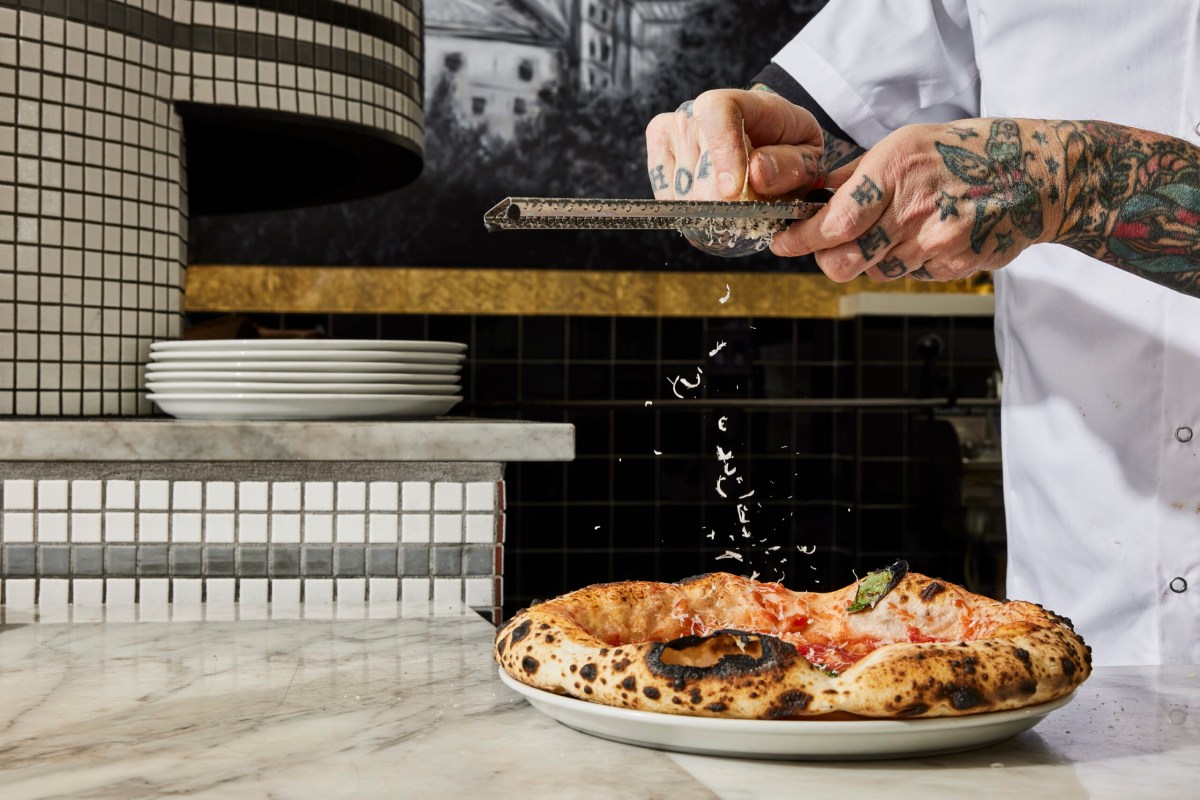 Chef's tattooed hands grating cheese over a freshly baked Neapolitan pizza on a marble counter, pizza oven behind.